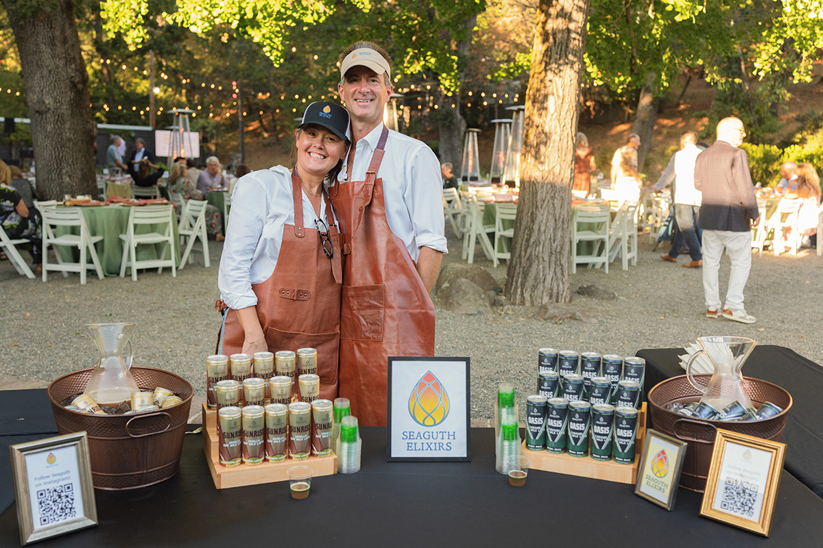 A couple stands beside a table displaying several bottles of wine, smiling and enjoying each other's company.