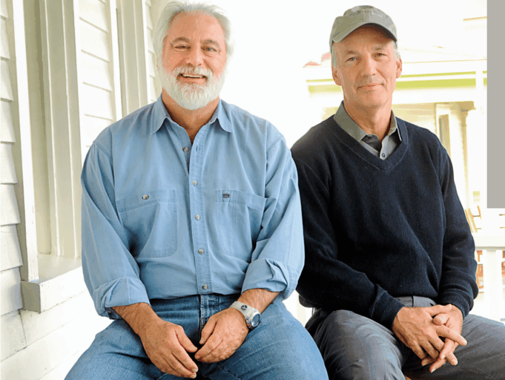Two older men are sitting on wooden benches outside on a porch, their smiles hinting at an amicable meeting of minds. The man on the left has a white beard and wears a light blue shirt with jeans, while the man on the right sports a grey cap, dark sweater and grey pants.