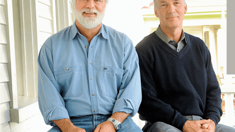 Two older men are sitting on wooden benches outside on a porch, their smiles hinting at an amicable meeting of minds. The man on the left has a white beard and wears a light blue shirt with jeans, while the man on the right sports a grey cap, dark sweater and grey pants.
