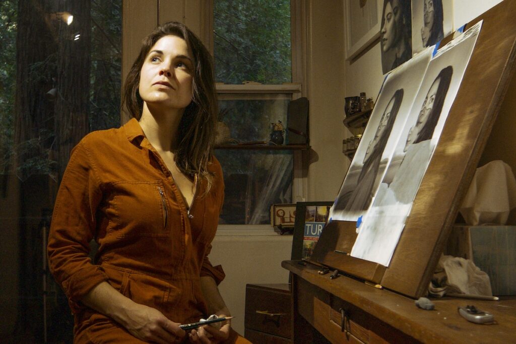 A woman wearing a copper colored shirt stares off into the distance in front of an easel with her art.