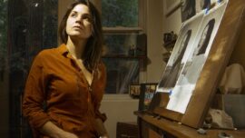 A woman wearing a copper colored shirt stares off into the distance in front of an easel with her art.