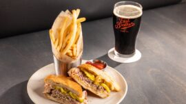 A plate with a sandwich sliced in half, a side of fries and a beer with the East Brother Beer Co. logo.