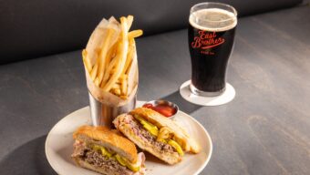 A plate with a sandwich sliced in half, a side of fries and a beer with the East Brother Beer Co. logo.