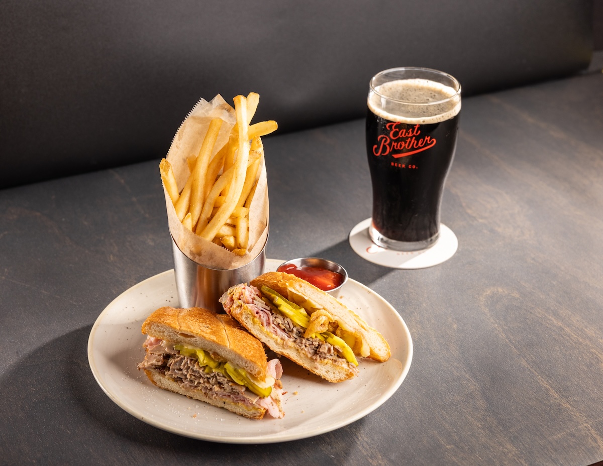 A plate with a sandwich sliced in half, a side of fries and a beer with the East Brother Beer Co. logo.