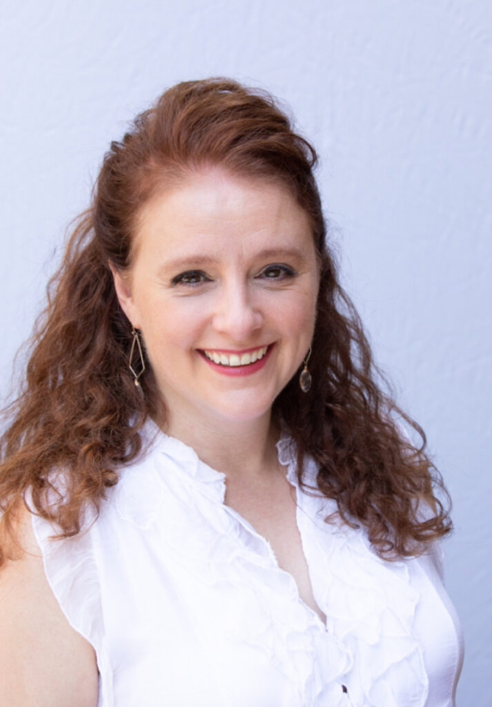 A headshot of a woman wearing a white tank top blouse and red curly hair.