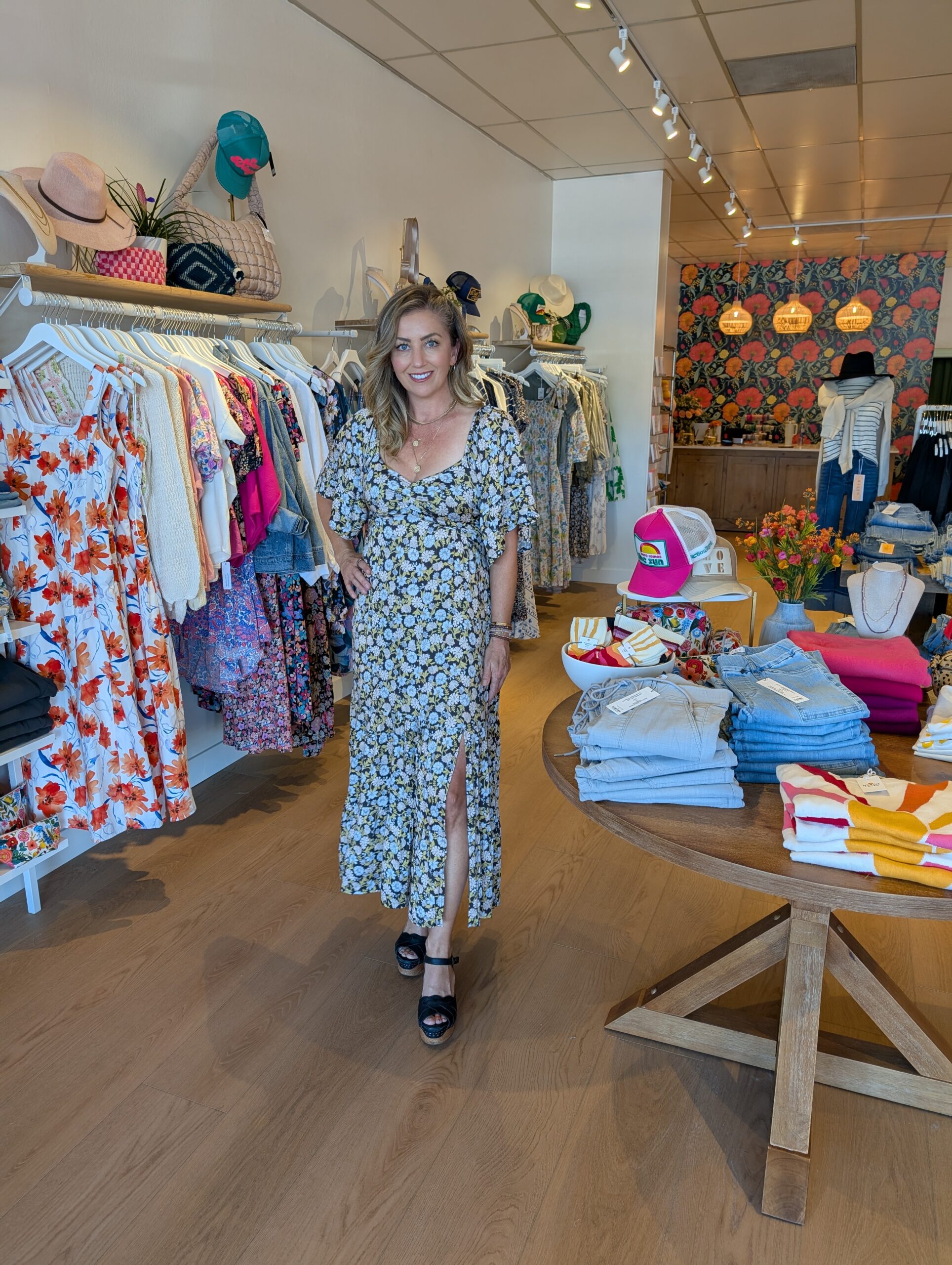 A woman wearing a long flowy dress poses in a shop with clothing and knick knacks.