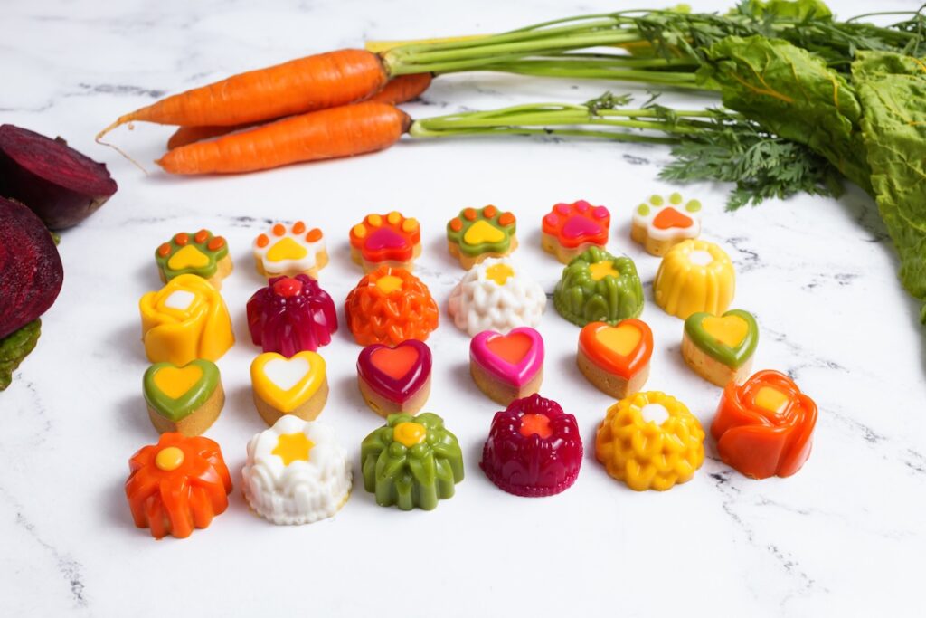 A white marble countertop with colorful gourmet dog treats and a bunch of carrots.