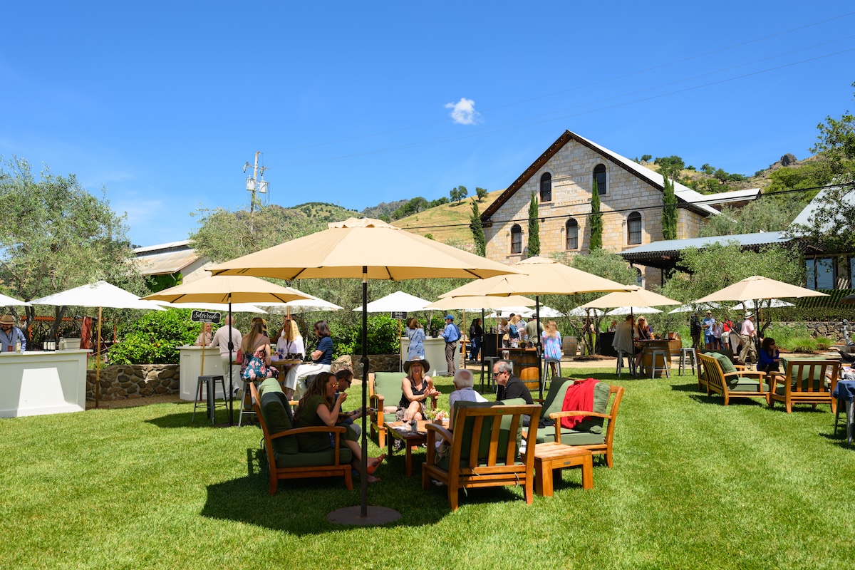 A green field with lawn chairs in front of a stone-brick winery, with people drinking wine in the chairs