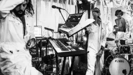 Black and white photo of a band consisting of keyboard player, singer and drums, playing in front of Marilyn Monroe poster
