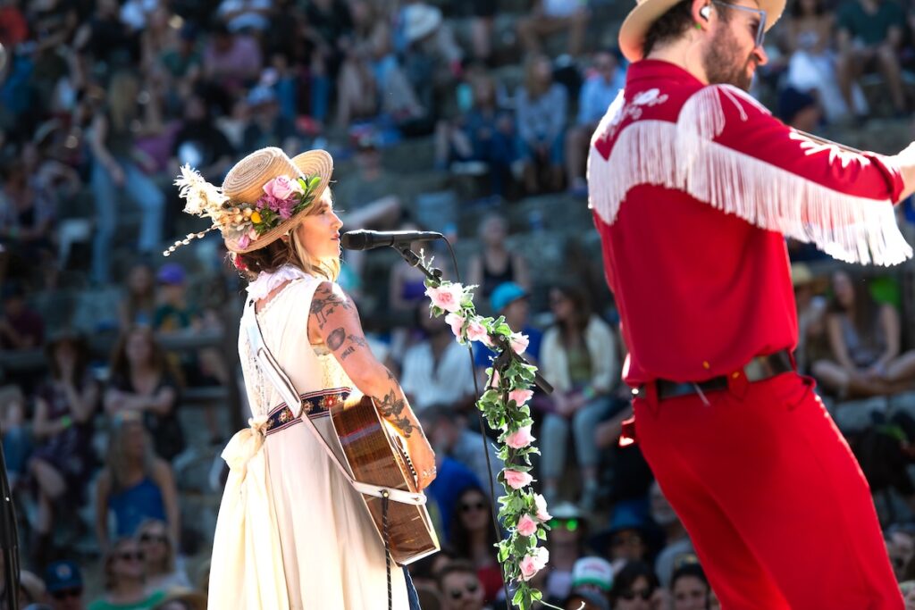A woman in a white dress plays guitar and sings on stage next to a man in a red outfit.