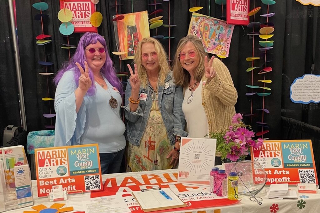 Three women stand behind a colorful booth for Marin Arts.