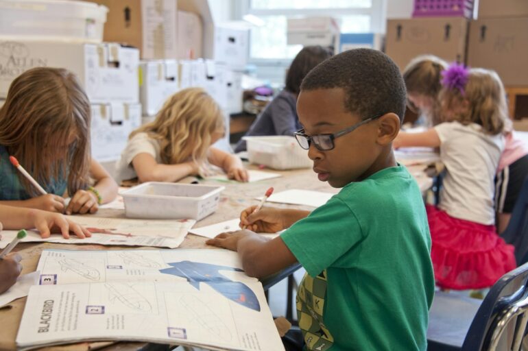 Kids sit at a table in a classroom and are reading and writing.