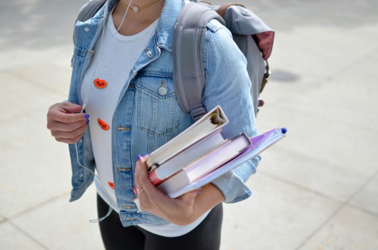 A girl wearing a backpack and holding an armful of textbooks.