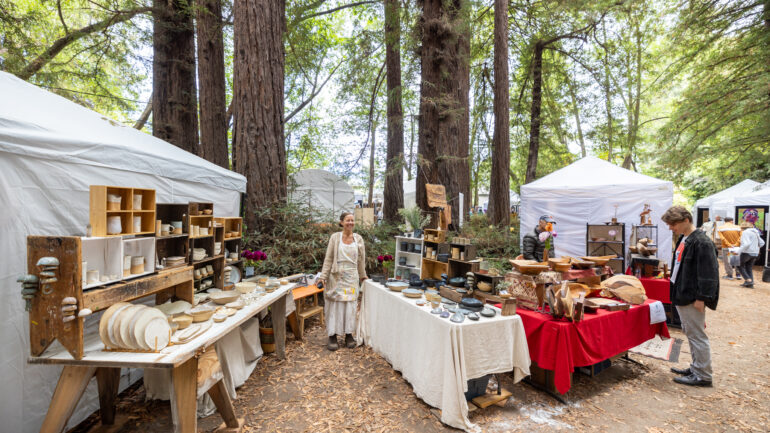 2024 MV Fall Arts Festival, photo of a guest shopping at a couple of the booths, with the trees in the background.