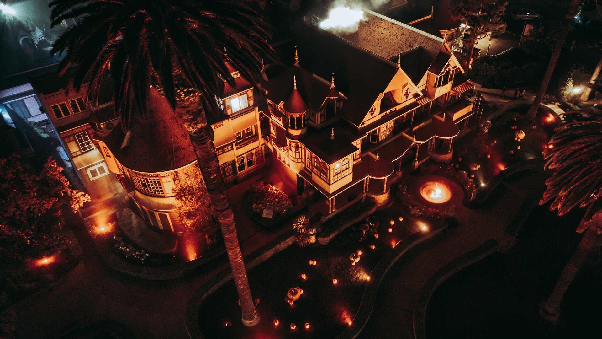 Bird's eye view shot of the Winchester Mystery House during the Halloween season. It's night and the house and lawn is lit by orange lighting.