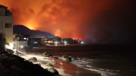 The bright orange flames of a wildfire lights up a hill from behind, with the view of a city and a beach in front.