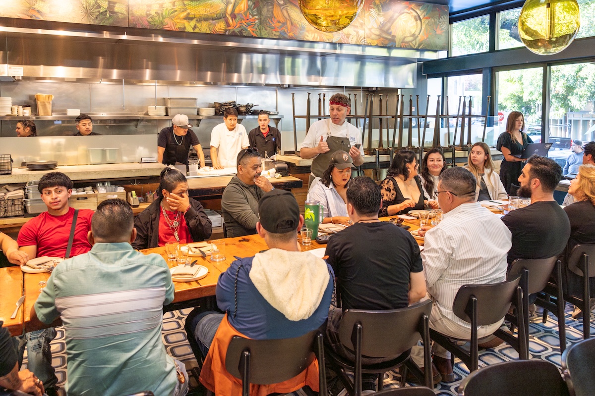 A large group of people sits at a long table at a restaurant with a view of the kitchen in the background.