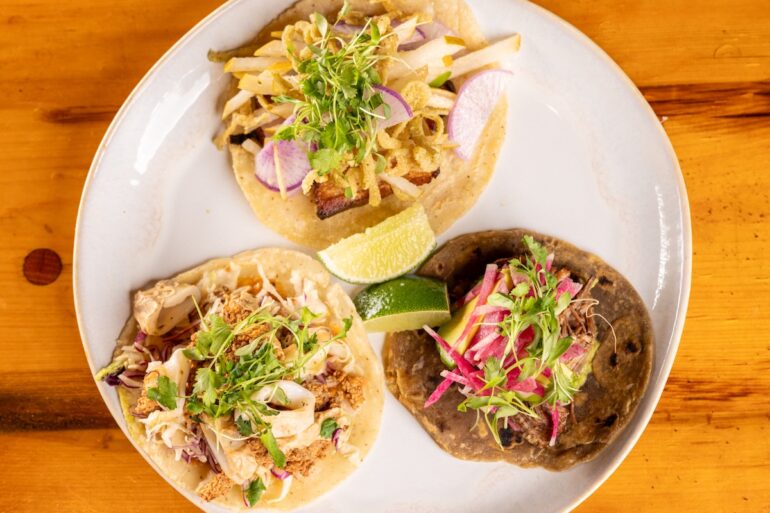 Three tacos sit on a plate with flour tortillas and a darker tortilla, with various toppings and lime for garnish.