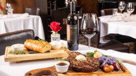 A wooden board set with a cut of meat, purple cauliflower and a dipping sauce; another board with bread; and a bottle of wine and wine glass on a table with a white tablecloth.
