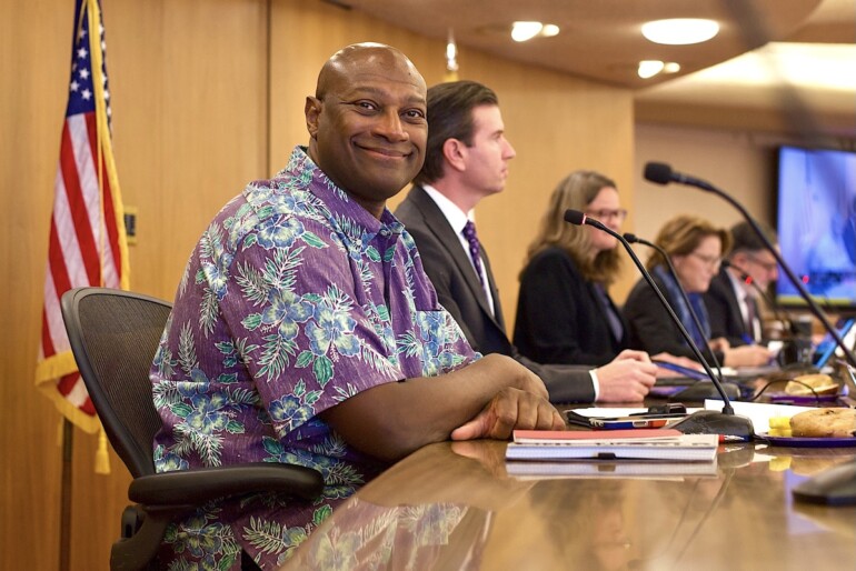 Brian Colbert Sits at a table in front of a microphone and notebook wearing a purple Hawaiian shirt as he's sworn in.