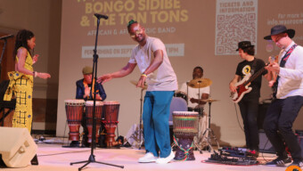 A man standing in front of a microphone on a stage dances while his band of five others play their instruments behind him.