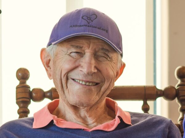 Jerry Jampolsky smiles, wearing a purple baseball cap and indigo sweater with a pink collar sticking out.