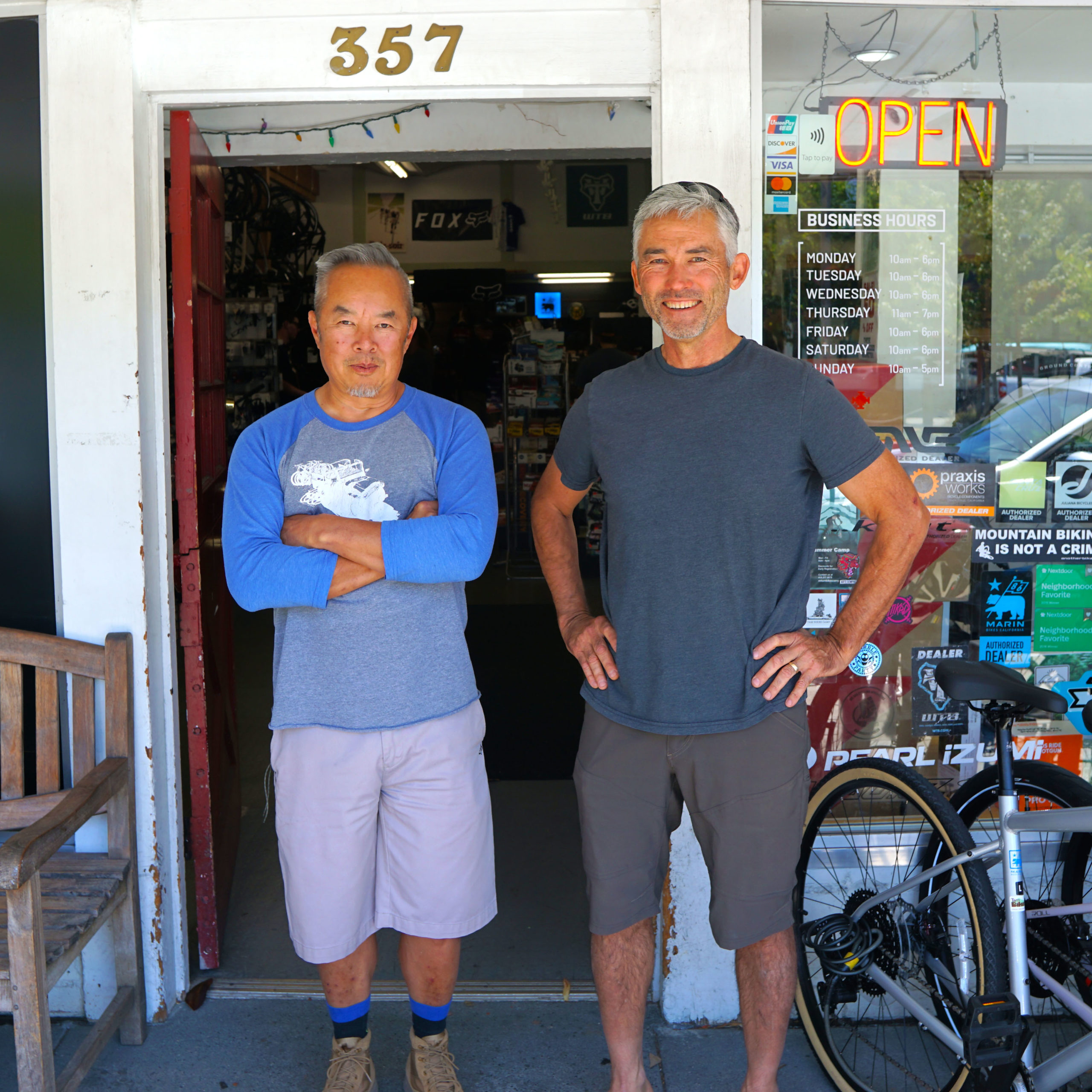 Two men stand in front of a bike shop with a sign that says "Open."