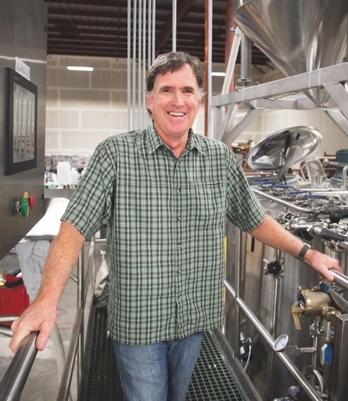 Chris Coomber wears a plaid shirt and stands in front of the machinery that brews beer. Vertical option.