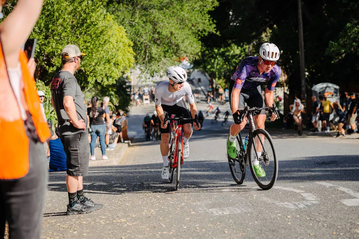 Two men on bikes are depicted going uphill in a race, with lots of people standing on the sides.
