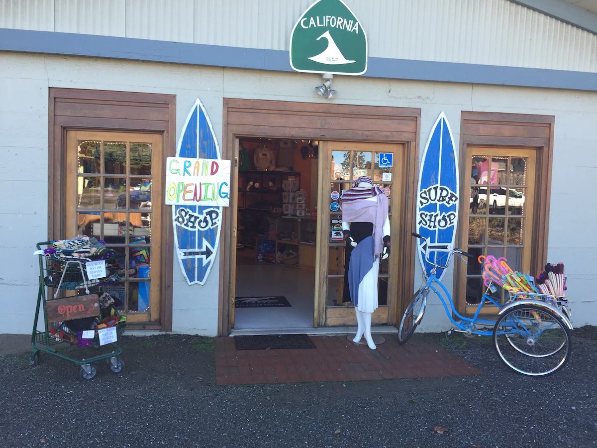 An exterior shot of a surf shop, with two signs in the shapes of surf boards, a shopping cart displaying merchandise and a bike all sitting out front.