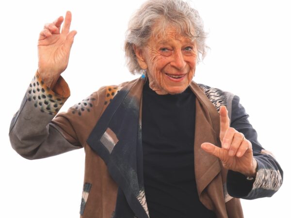 Anna Halprin wears a patterned coat and dances with her arms up.
