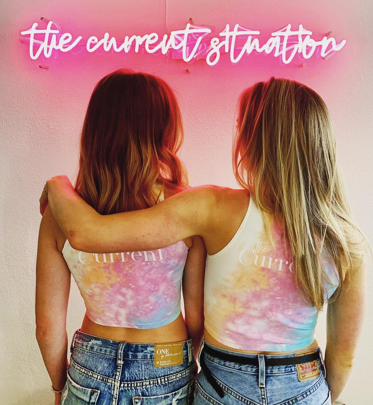 One woman with arm over another woman, neon sign saying "The Current Situation."