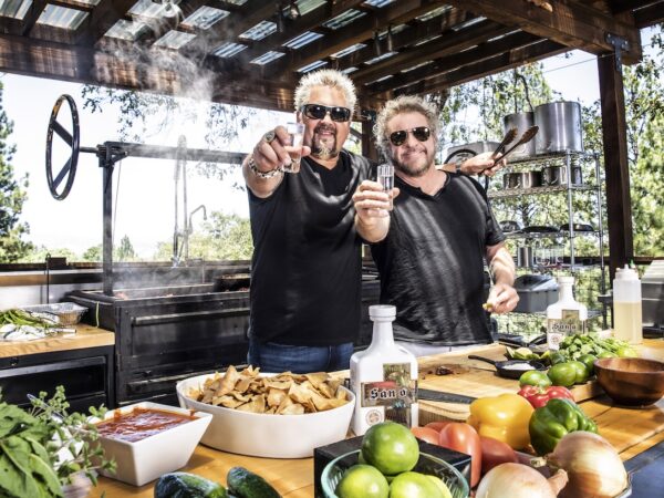 Guy Fieri and Sammy Hagar hold up shot glasses in the midst of an outdoor kitchen.