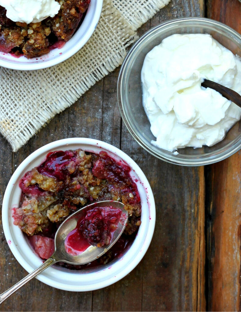 A small white ramekin filled with berry and rhubarb crisp, topped with a golden oat crumble and served with a spoon. A bowl of whipped cream sits nearby on a rustic wooden table with burlap fabric.