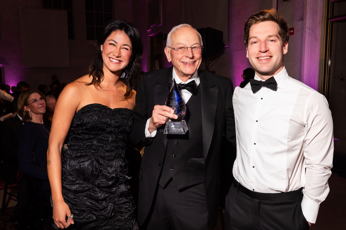Two men wearing tuxedos standing with a woman in a black, long strapless gown.