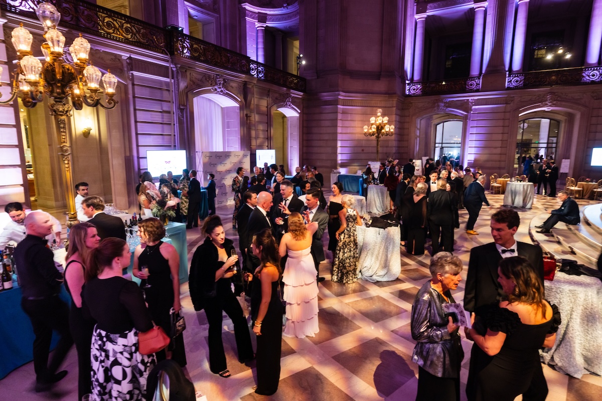 A large group of people in cocktail attire chat on a dance floor lit with purple lights.
