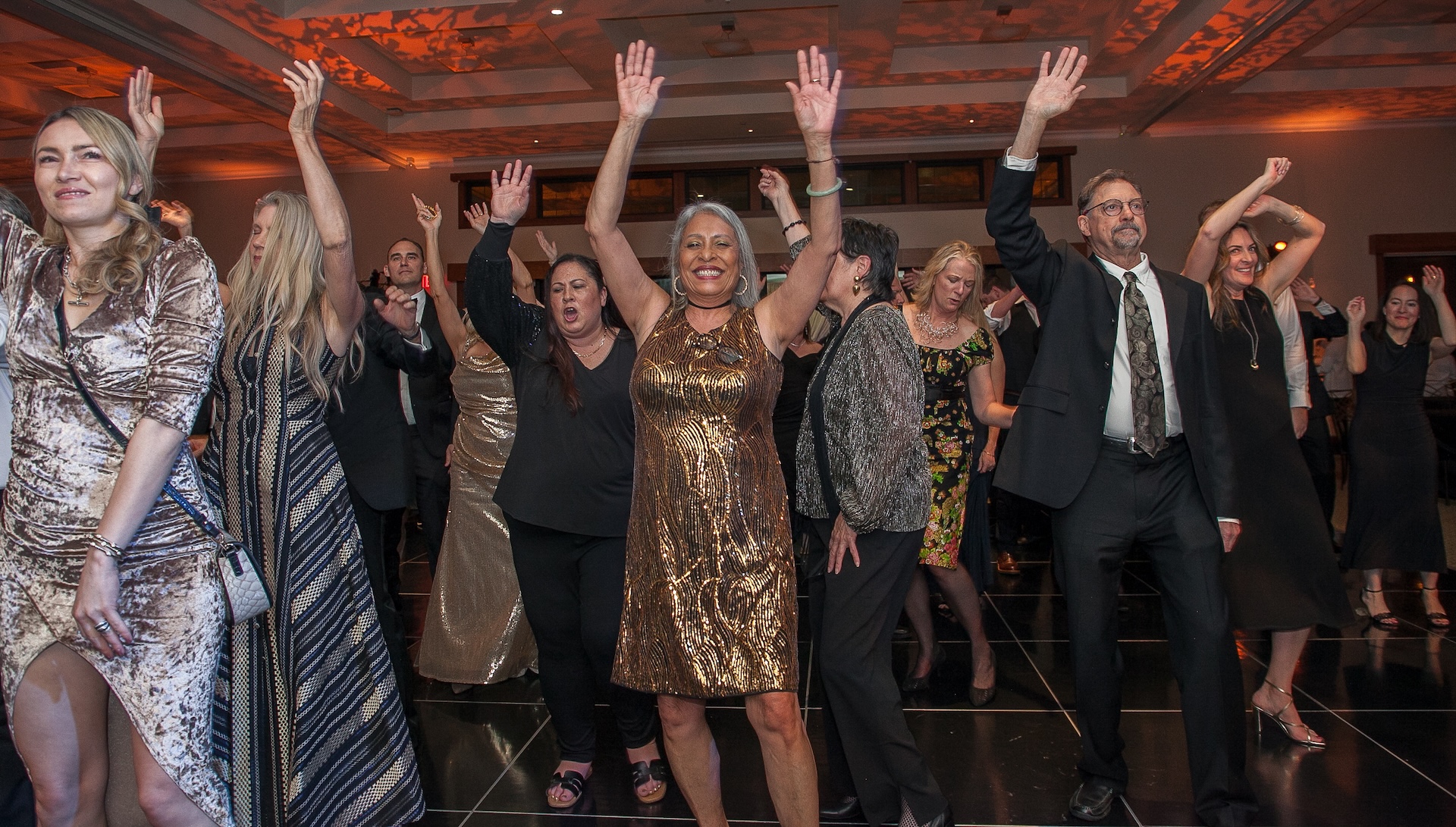 A group of people in cocktail attire dance on a dance floor with their hands raised.