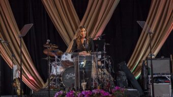 A woman wearing a black pantsuit sits at a drum set center stage.