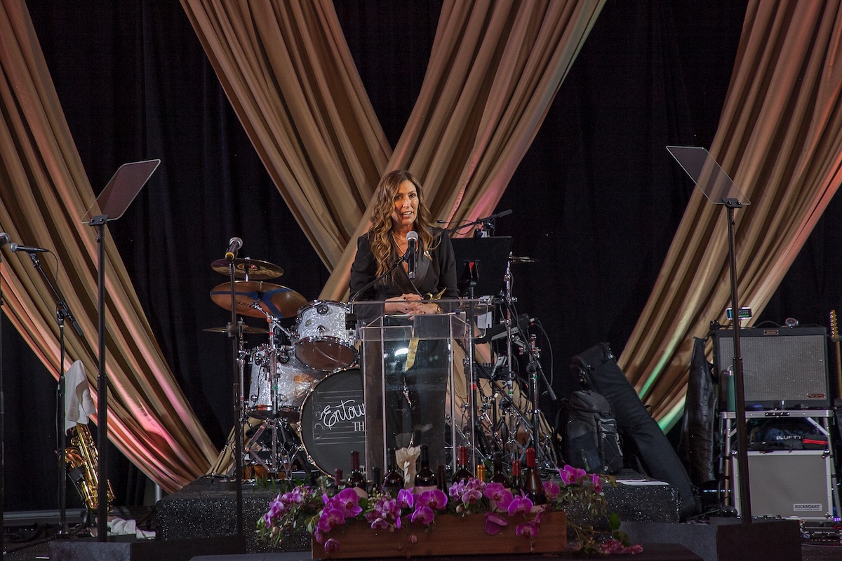 A woman wearing a black pantsuit sits at a drum set center stage.