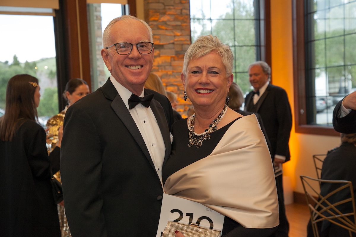 A woman with short silver hair wearing a black dress and gold shawl, standing with a man in a classic tuxedo.