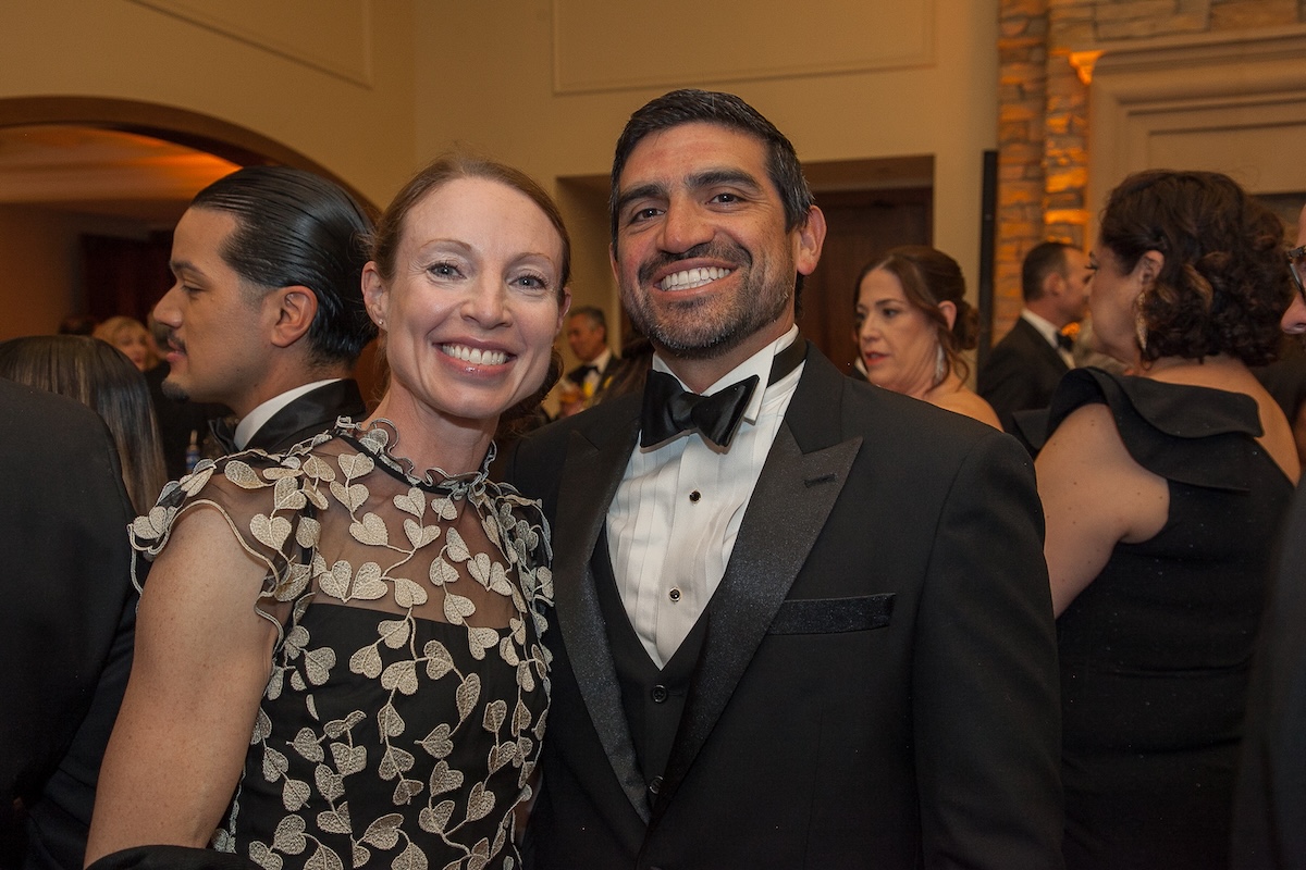 A man wearing a classic tuxedo stands with a woman wearing a black mesh dress with leaf patterning.