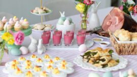 A spring brunch spread with deviled eggs, asparagus pastry bundles, biscuits, shot glasses with beet soup, mini cupcakes, flowers, decorative eggs and ceramic bunnies.