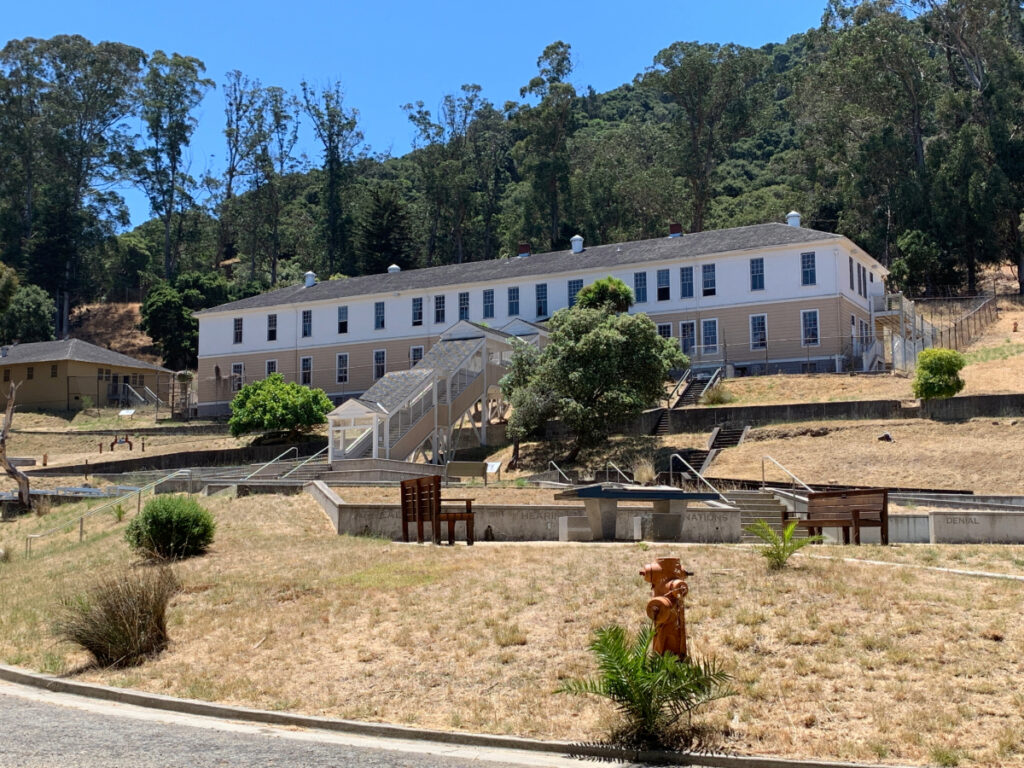 A wide view of large white and beige wooden buildings set against a hill, once used for detaining immigrants.