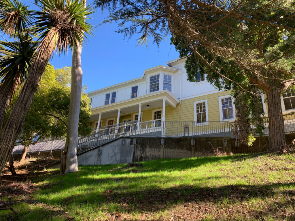 A historic yellow and white building surrounded by trees and grass under a clear blue sky, once a hospital and now a museum.