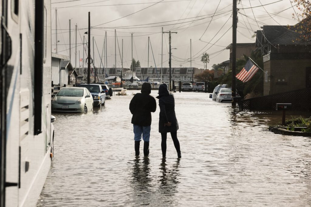 Two people stand in knee-deep floodwater on a residential street in Marin, surrounded by parked cars and boats in the distance, under a cloudy sky.