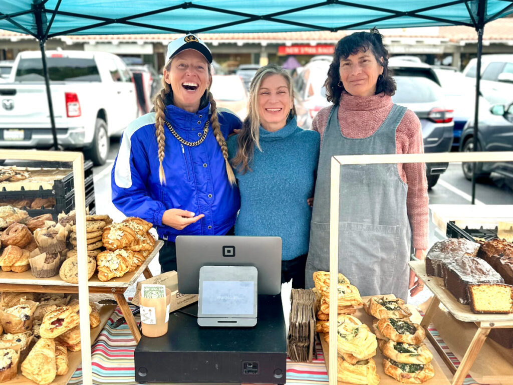 Three smiling women from Waldscraft Bakery pose behind a colorful farmers market stand filled with croissants, cookies, pastries, and loaves of bread.