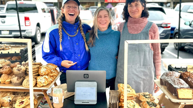 Three smiling women from Waldscraft Bakery pose behind a colorful farmers market stand filled with croissants, cookies, pastries, and loaves of bread.
