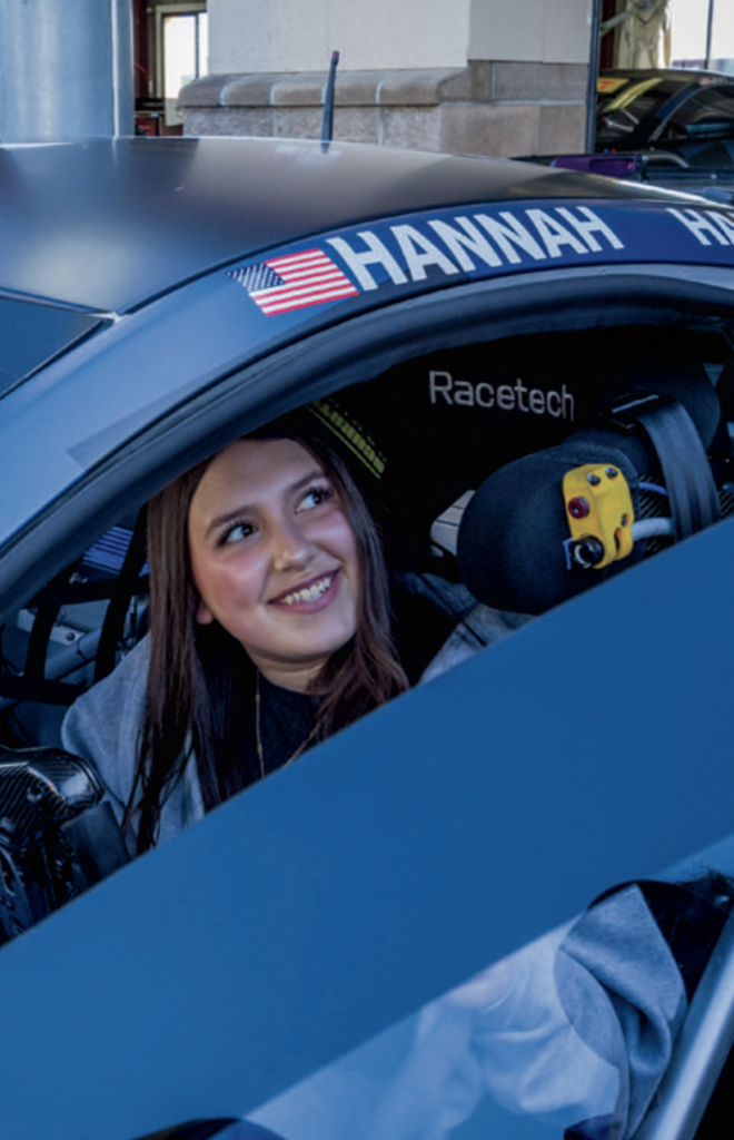 A young girl sits in the driver's seat of a race car and looks through the open window.