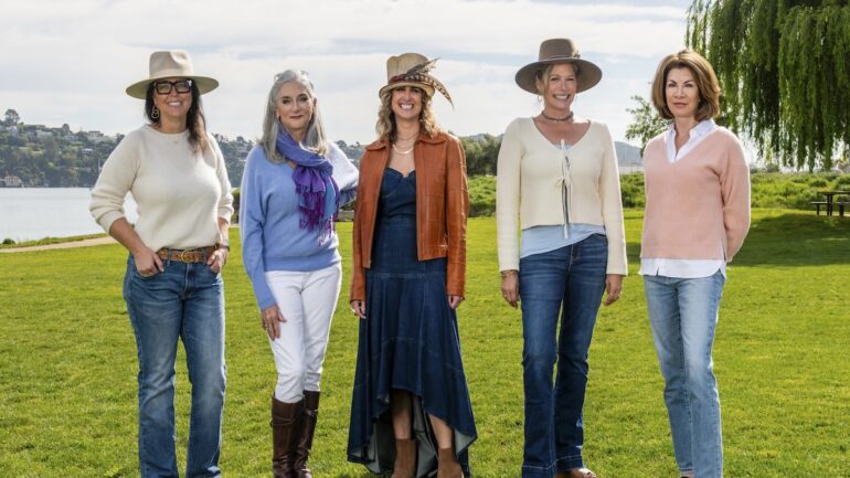 Five stylish women standing on a grassy field near the water under a sunny sky.