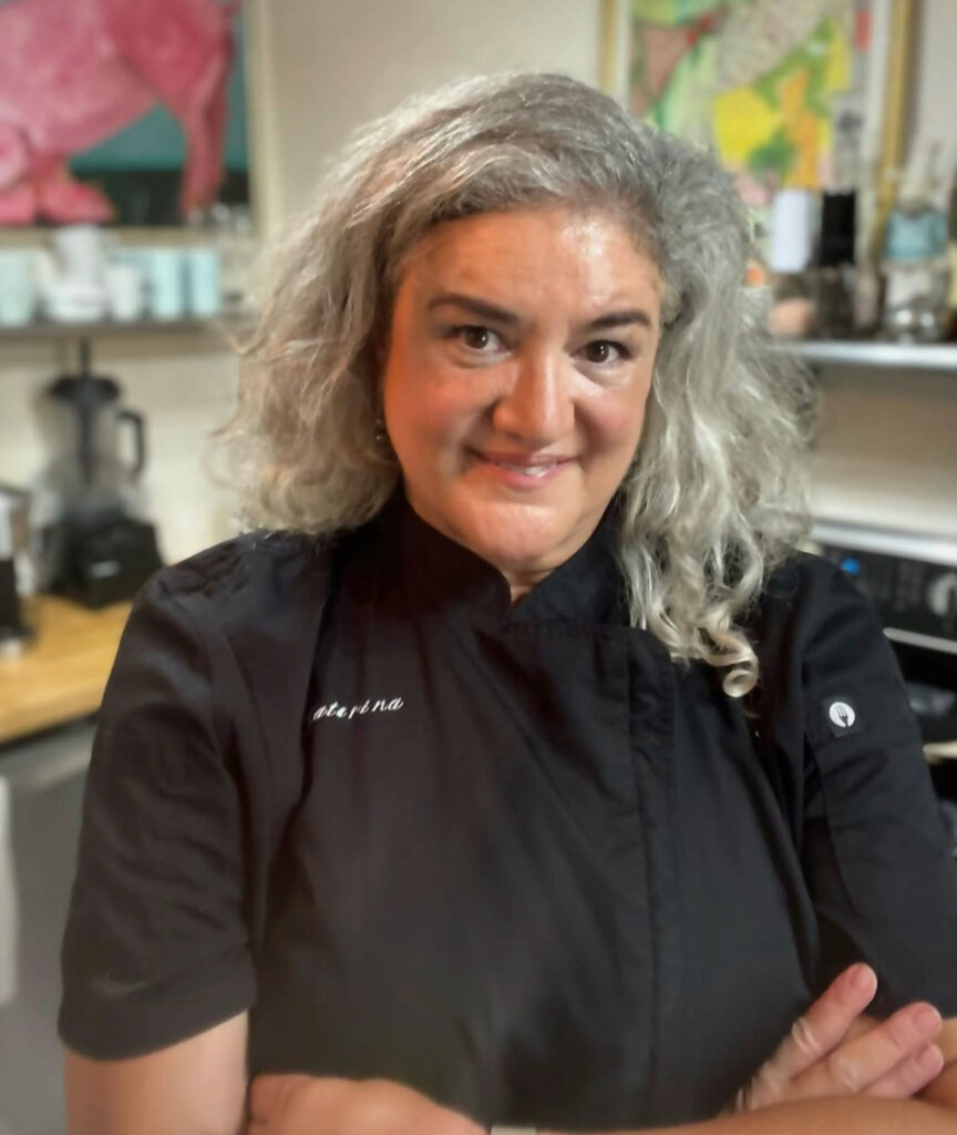 Chef Katarina Tualemoso standing in a kitchen, wearing a black chef’s jacket with her name embroidered on it, smiling with arms crossed.
